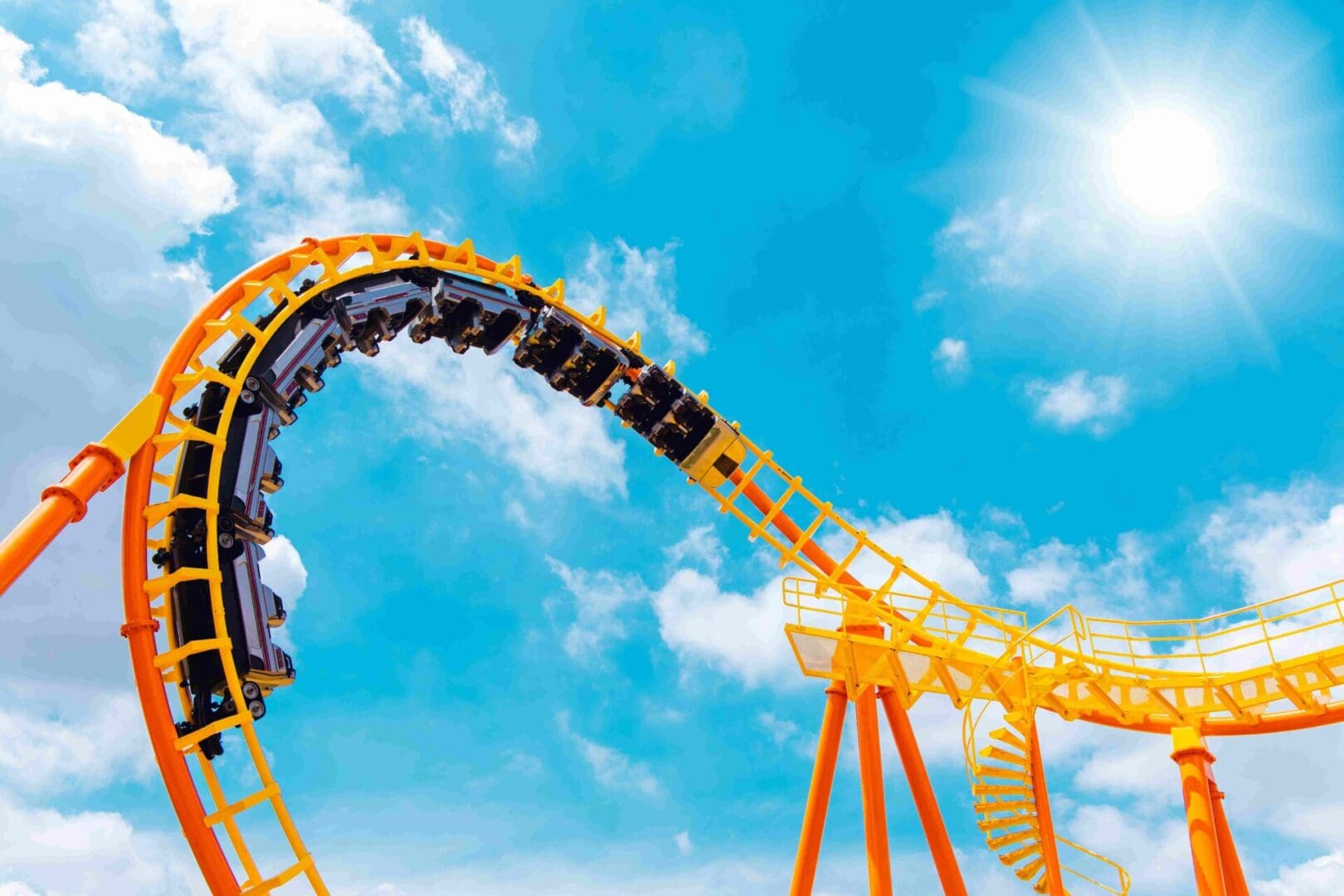 A yellow roller coaster with passengers upside down on a loop under a bright blue sky with the sun shining.