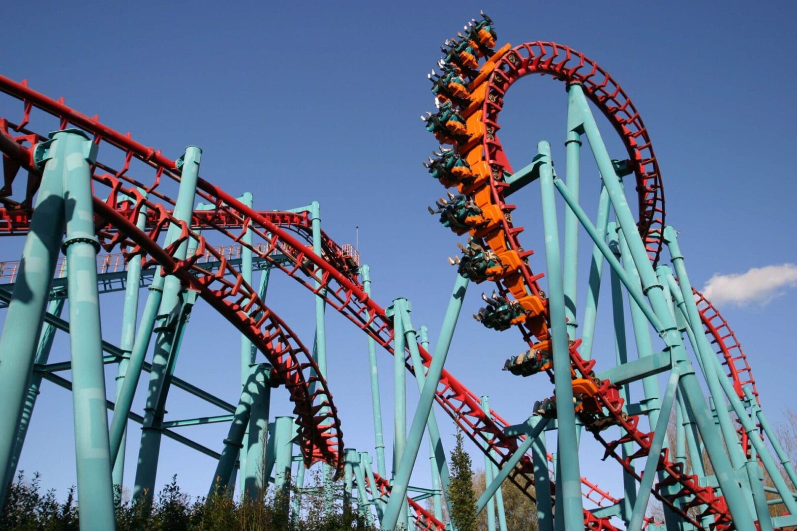 A red and teal roller coaster with a loop section under a clear blue sky. Riders are visible on the looping track.