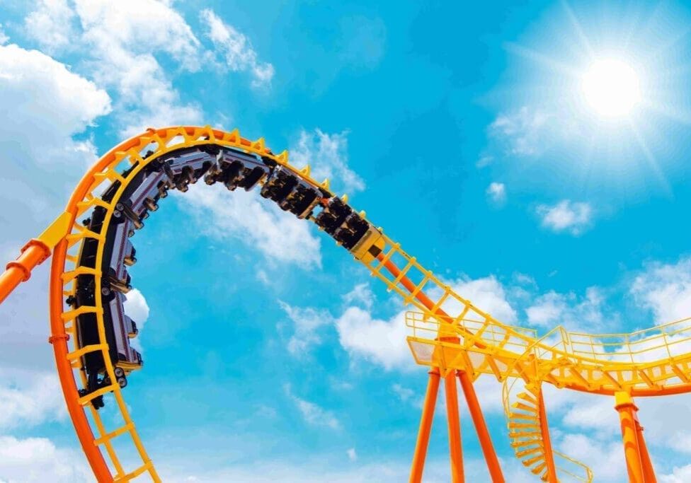 A yellow roller coaster with passengers upside down on a loop under a bright blue sky with the sun shining.