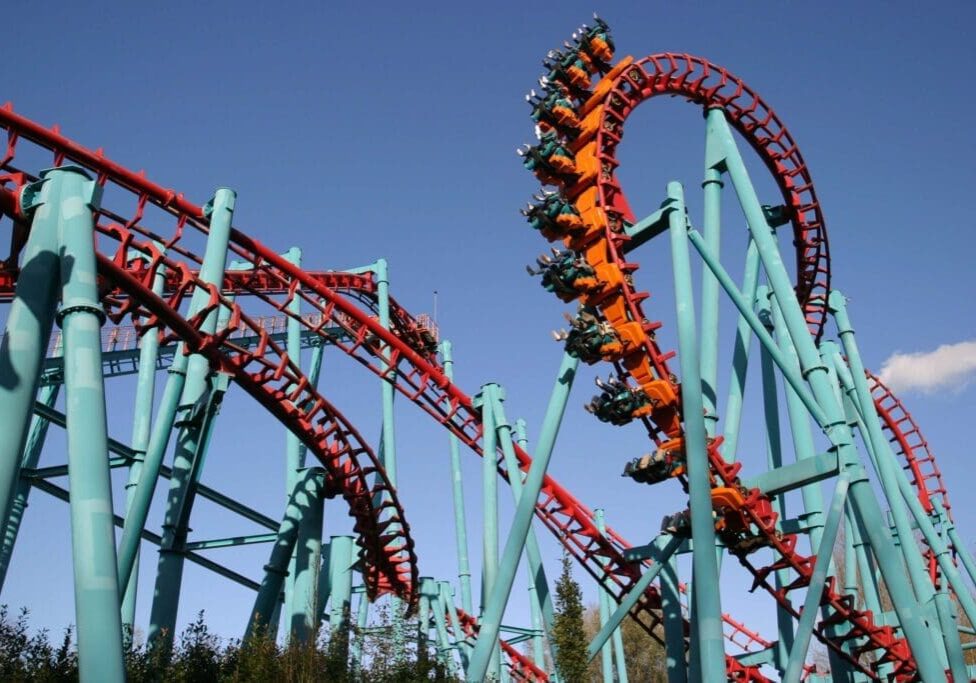 A red and teal roller coaster with a loop section under a clear blue sky. Riders are visible on the looping track.